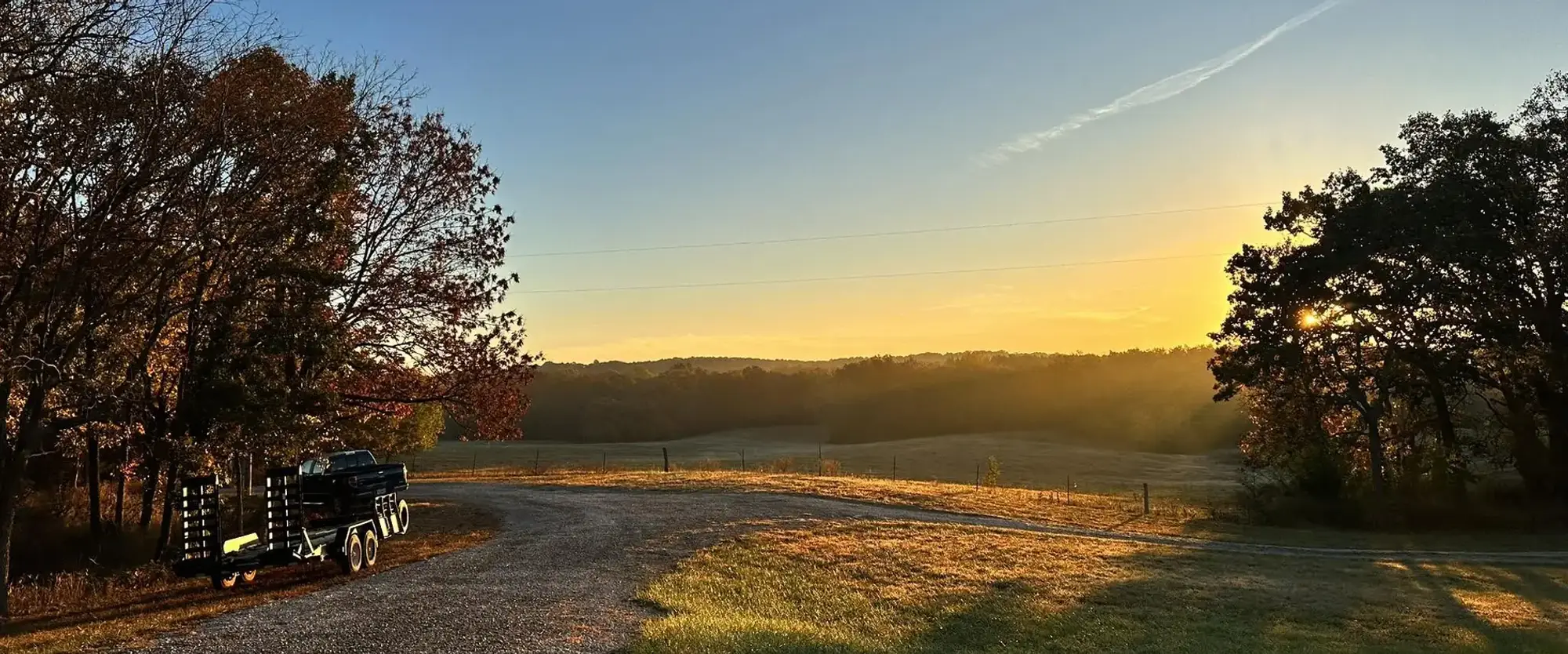 A gravel drive running out to a hayfield at sunrise, timber on both sides.